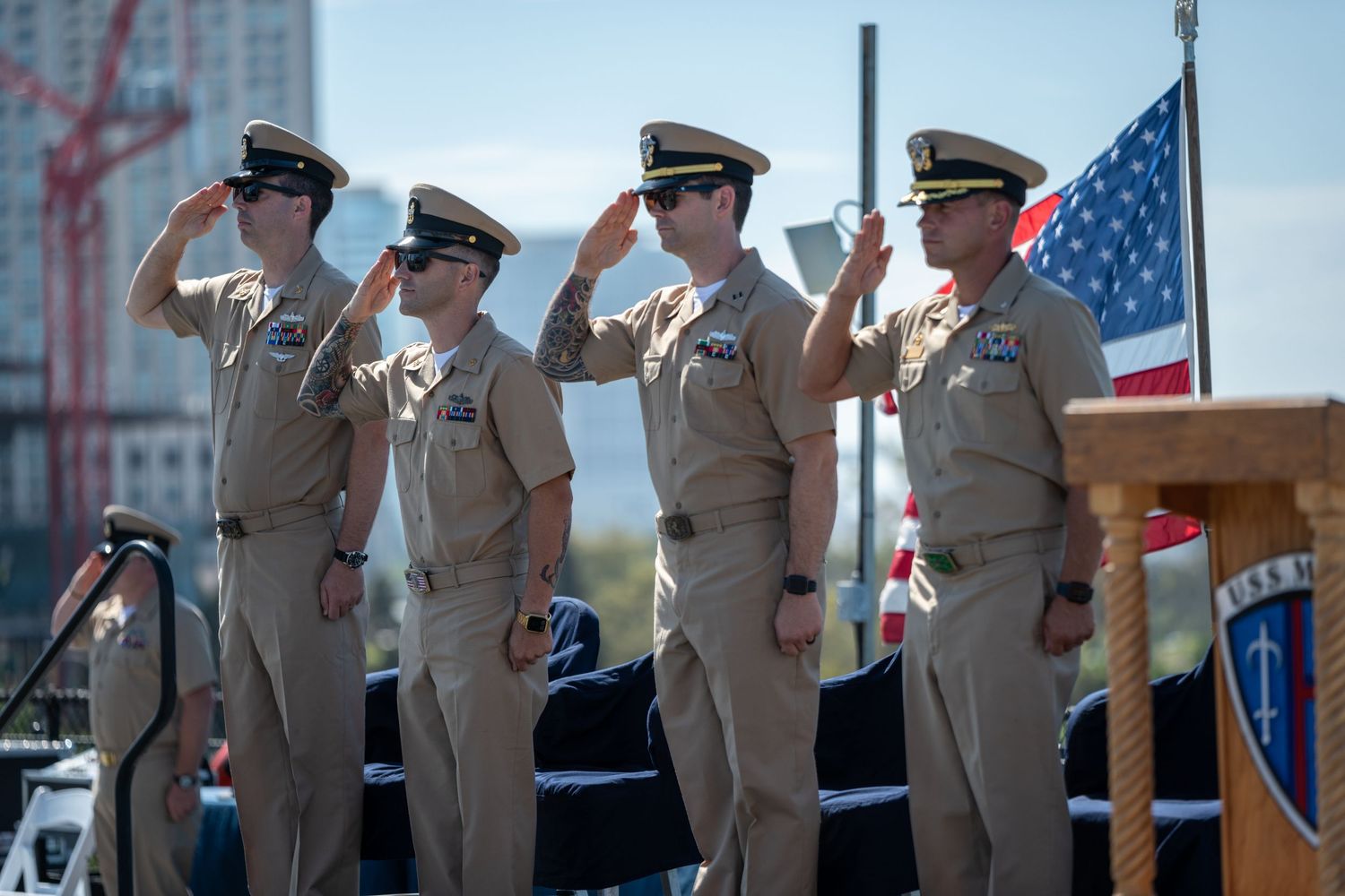 Military service members saluting in formation with American flag in background.