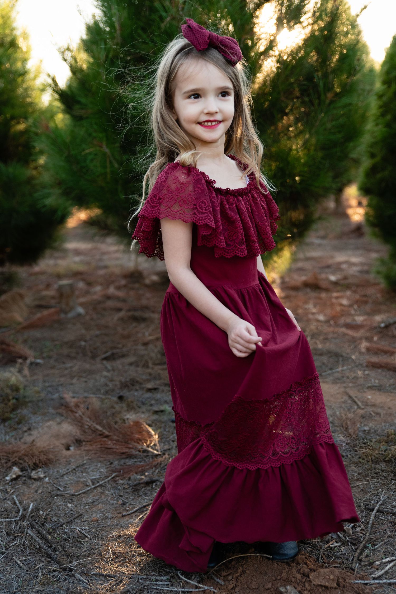 Young child twirls in a burgundy velvet dress near evergreen trees at sunset.