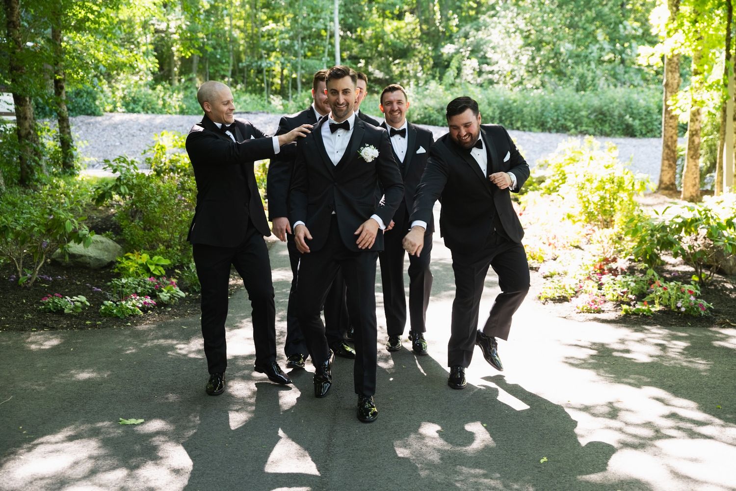 Groomsmen in black tuxedos jumping and posing playfully outdoors.