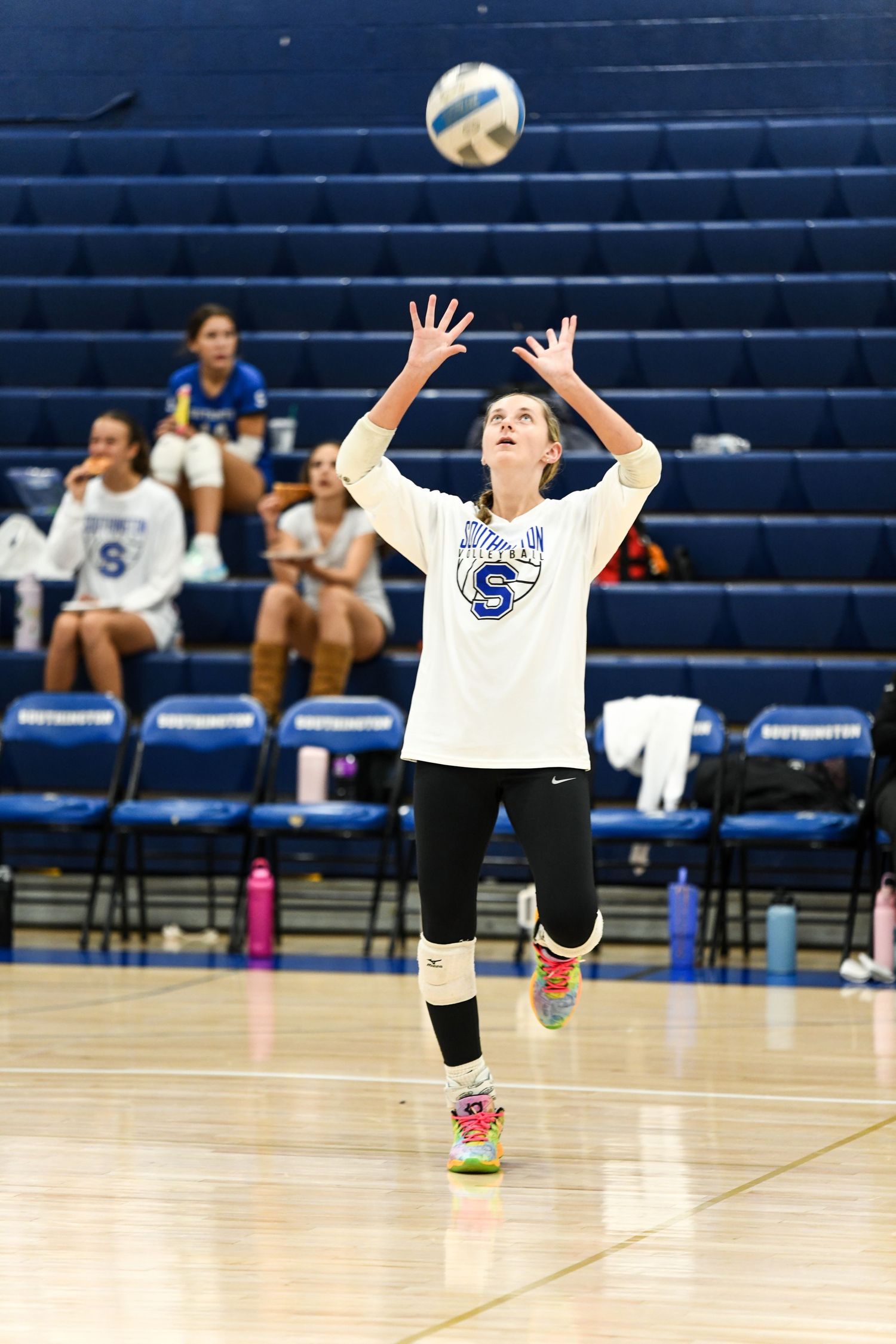 Female volleyball player in white jersey setting the ball during an indoor match with teammates on the bench.