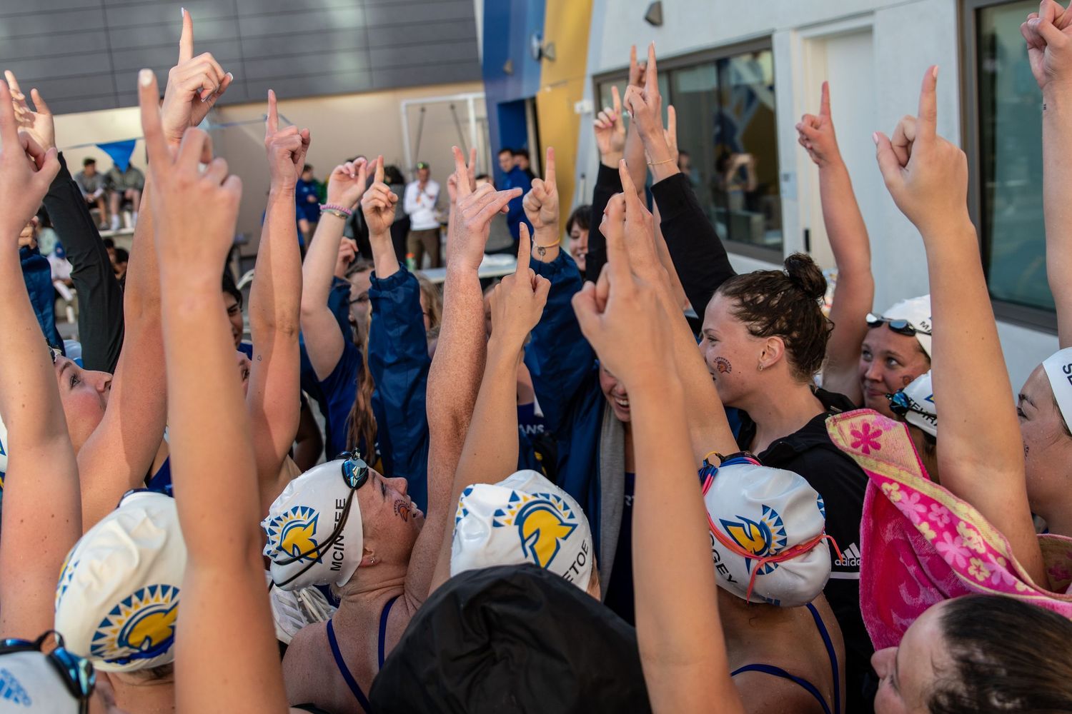 A swim team huddles together with raised hands in a team cheer at an aquatic event.