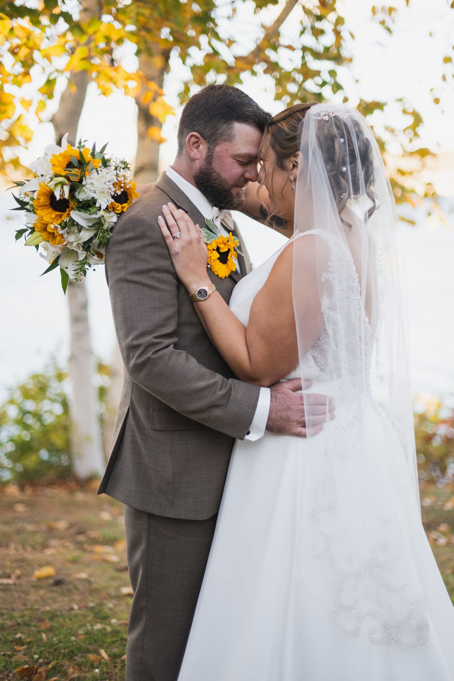 Autumn wedding couple shares emotional moment under yellow-leaved trees with sunflower bouquet.