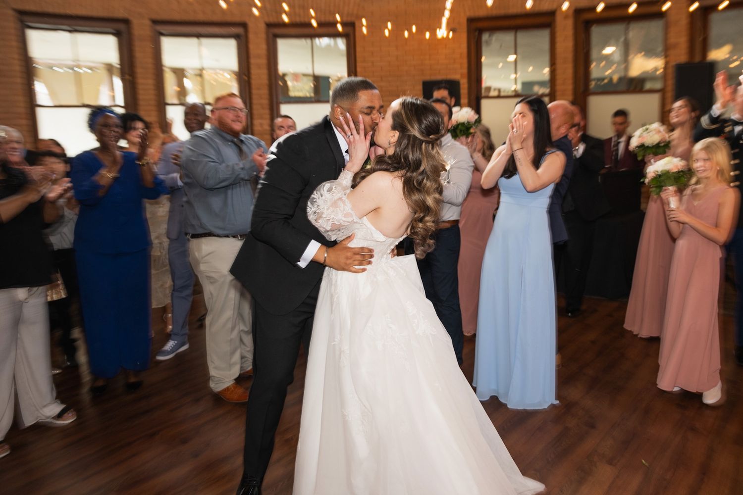 Wedding couple dances together at their reception under warm indoor lighting with guests watching.