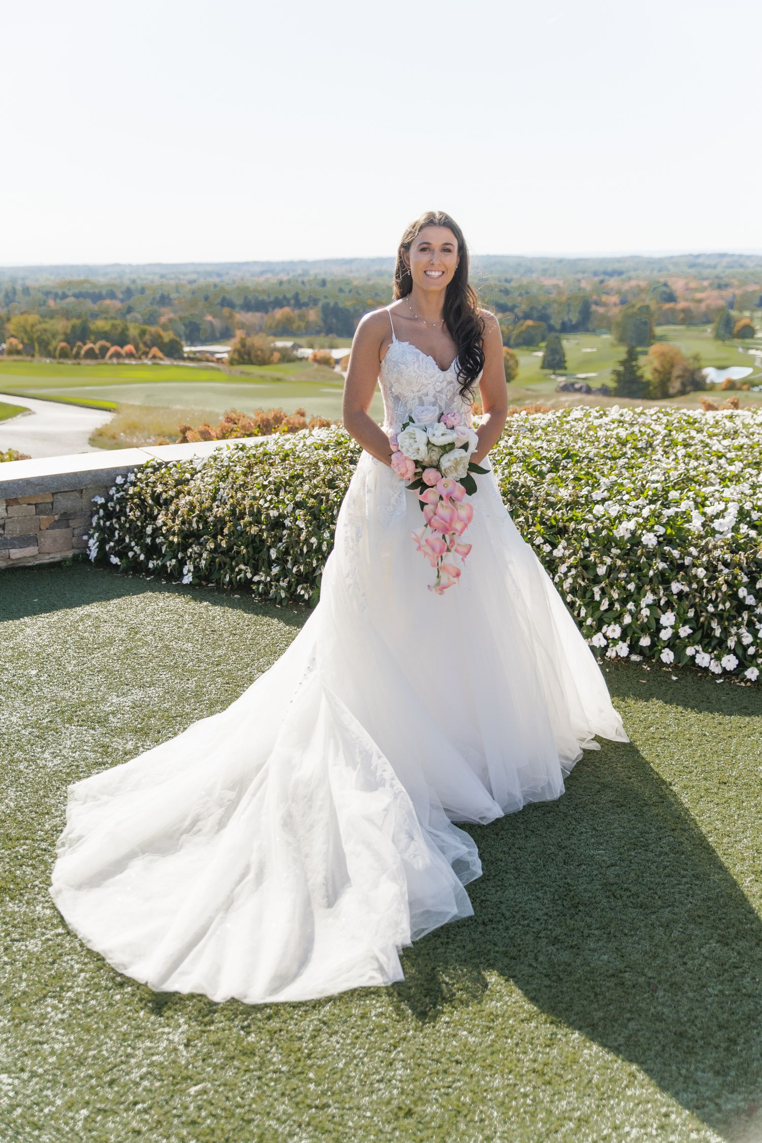 A bride in a flowing white wedding gown poses in a garden with white flowers and rolling hills in the background.