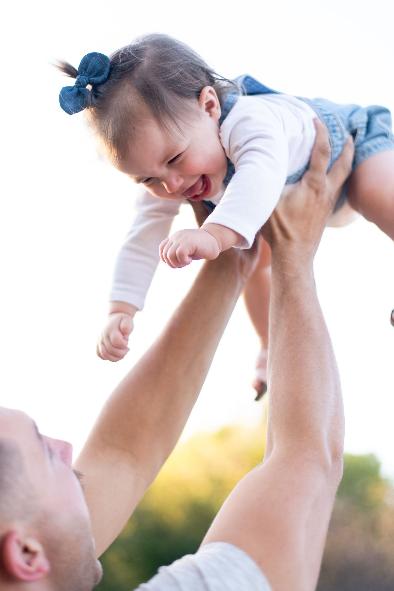 Parent lifting laughing baby wearing blue hair bow high in the air at sunset.