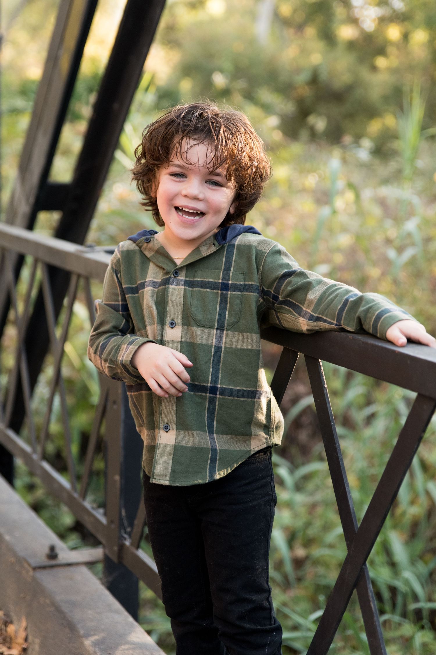 Playful child leaning on wooden bridge railing in plaid shirt.