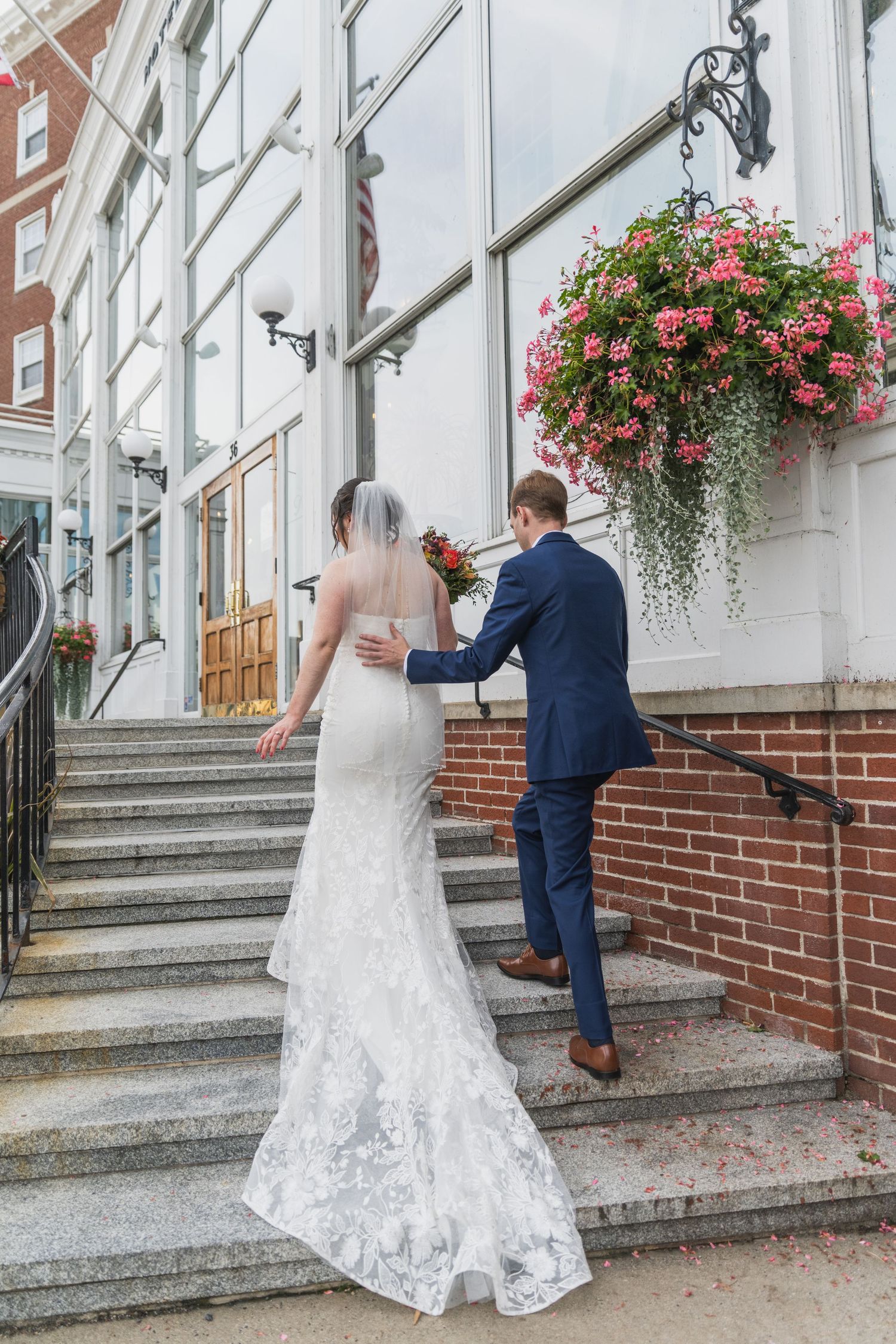 Couple in wedding attire ascends concrete steps with brick walls and hanging flower baskets.