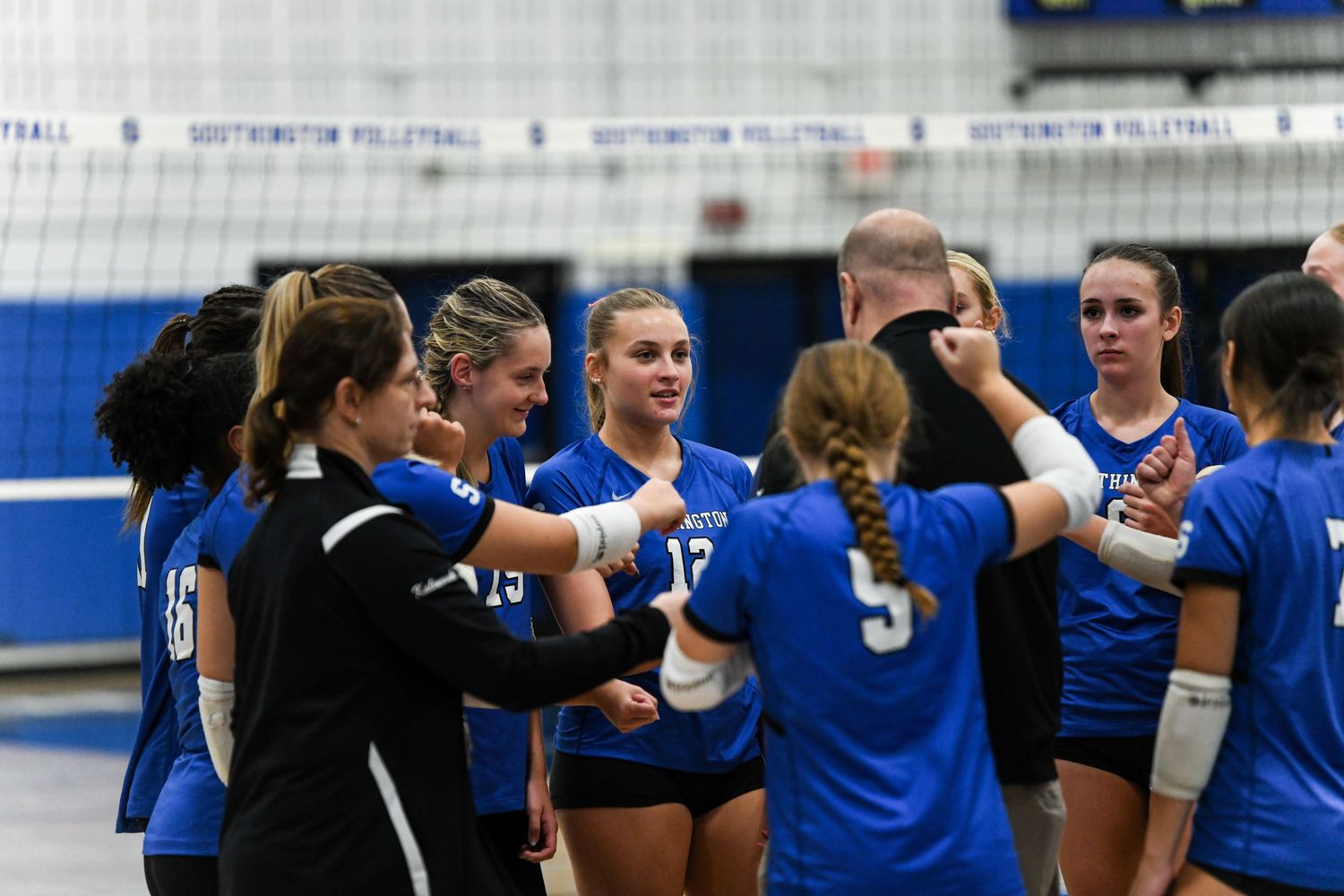 Southington volleyball team huddles together in blue uniforms during a match in a school gymnasium.
