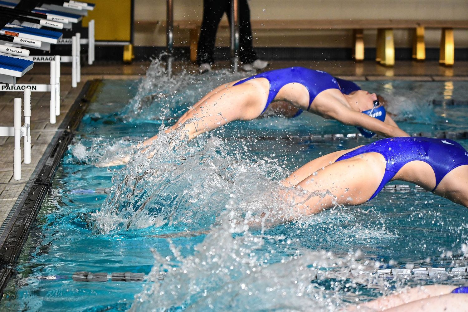 Two swimmers in blue suits dive into the pool at the start of a competitive swimming race.
