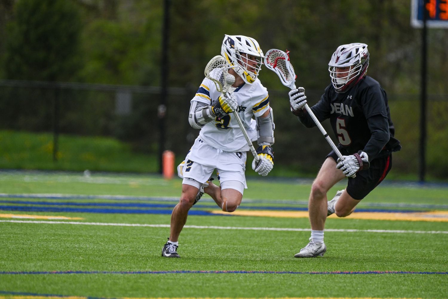 Two lacrosse players compete on a turf field, one in white and gold, the other in dark red uniforms.