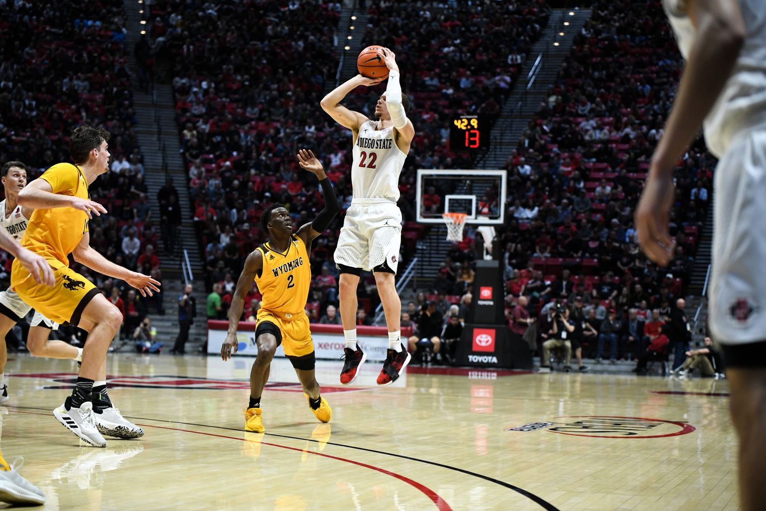 Basketball player #22 shoots a jump shot during a college game, with players in yellow and black uniforms defending.
