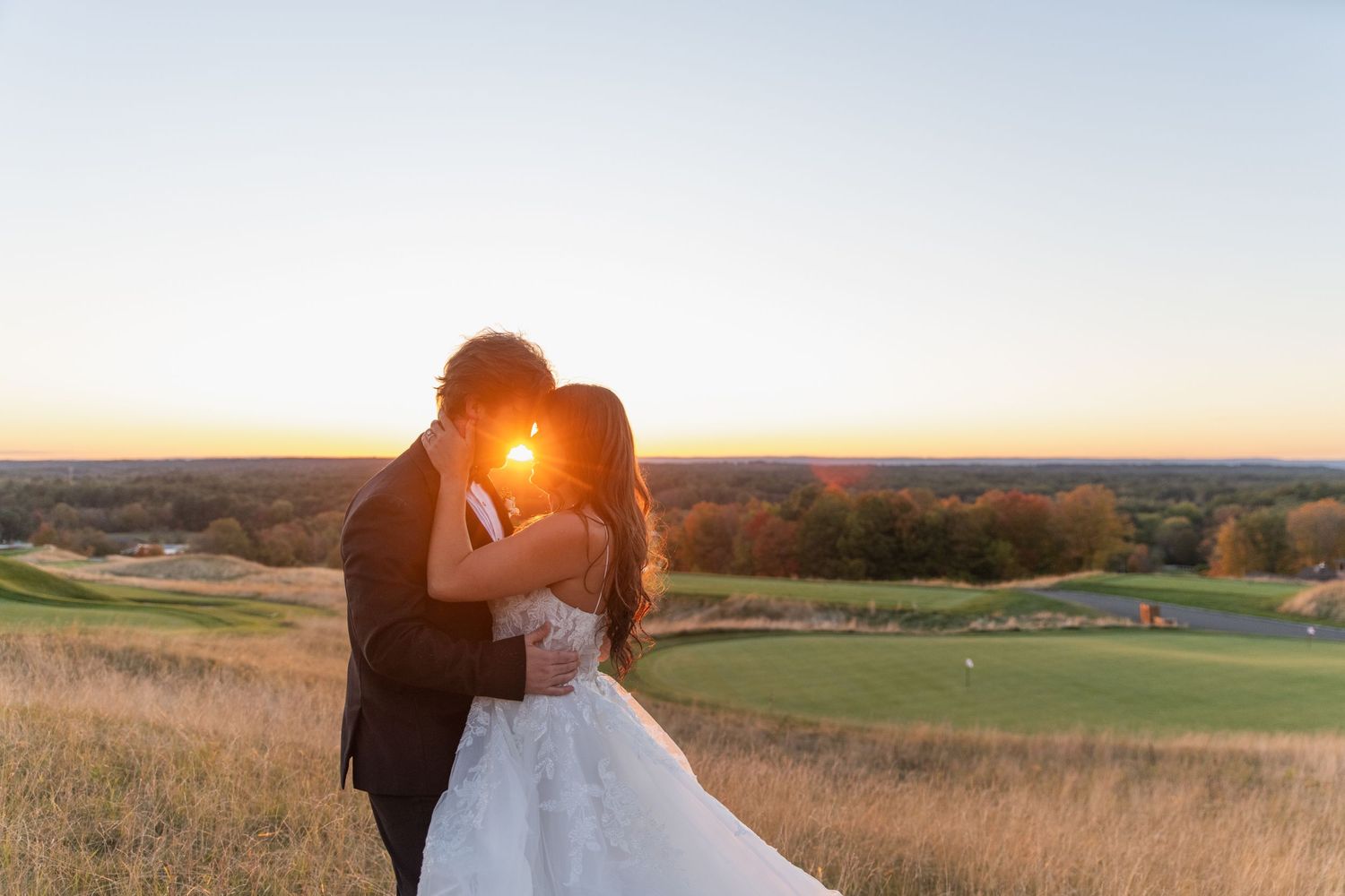 A couple in wedding attire shares an intimate embrace at sunset in a golden field with rolling hills in the background.