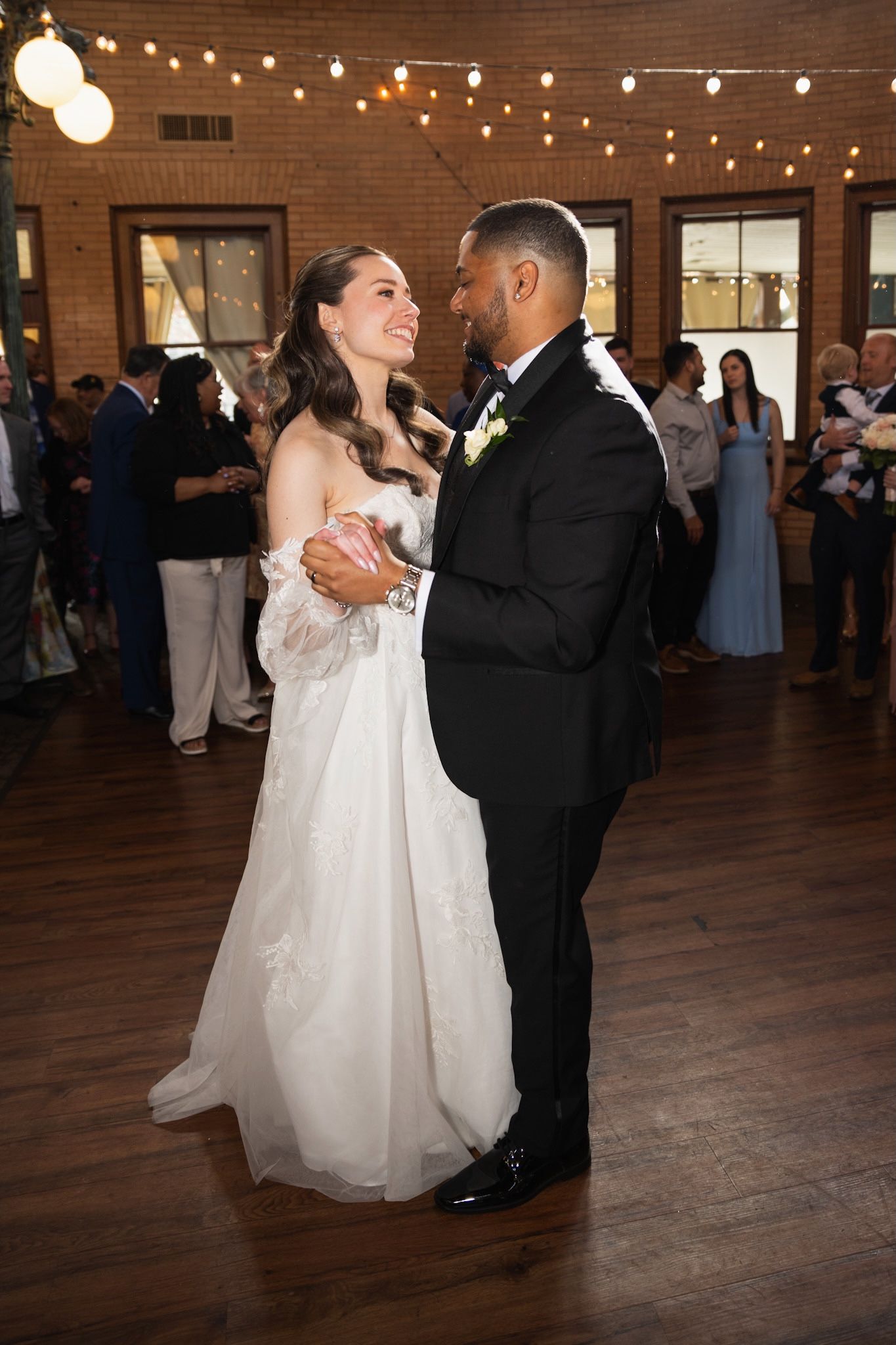 Wedding couple dancing at indoor reception with string lights and wooden floors.