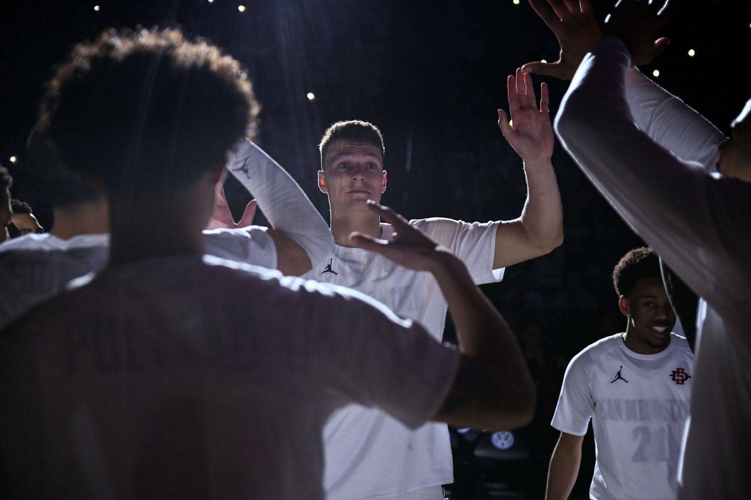 Basketball players in white Jordan Brand uniforms celebrate with high-fives during a pre-game huddle under dramatic lighting.