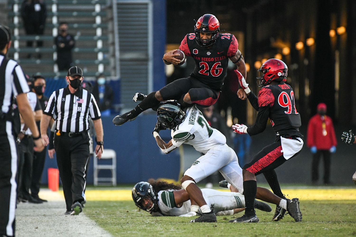 San Diego State football player #26 leaps over a Hawaii Warriors defender during a night game.