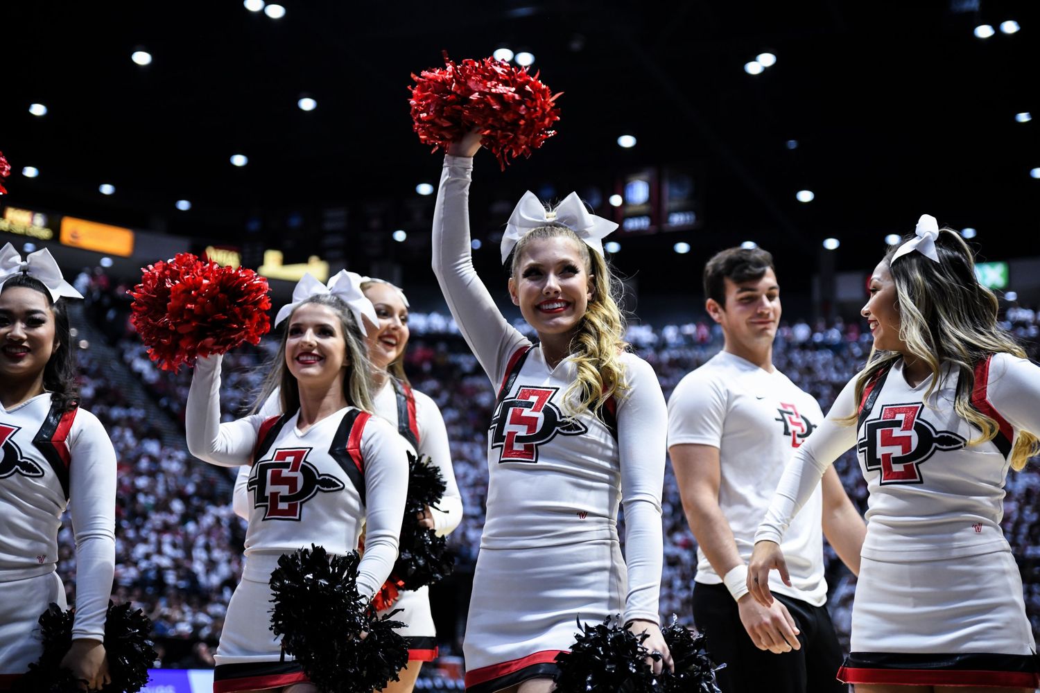 San Diego State cheerleaders in white uniforms wave red pom-poms during a college basketball game.