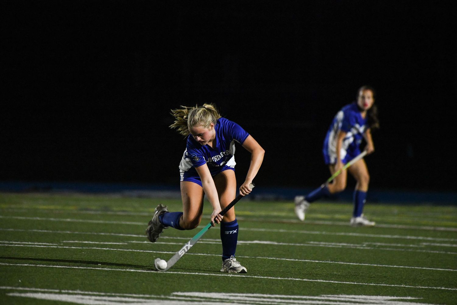 A female field hockey player in blue dribbles the ball during a nighttime game on a turf field.