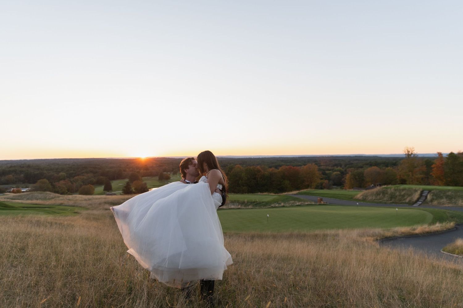 A bride in a flowing white dress walks through a golden field at sunset during a romantic outdoor photo session.