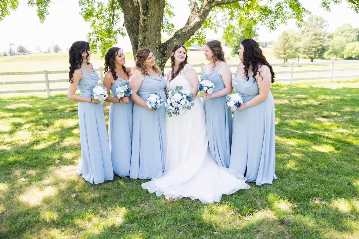 Bridal party in light blue dresses poses together under tree branches during outdoor wedding.