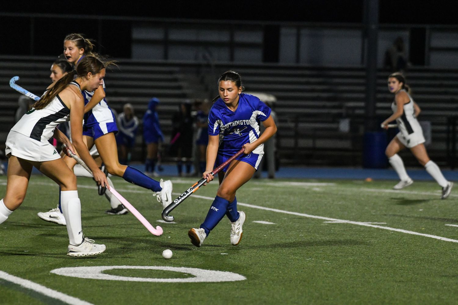 Female field hockey players compete on a lit turf field at night, with a player in blue dribbling past defenders.