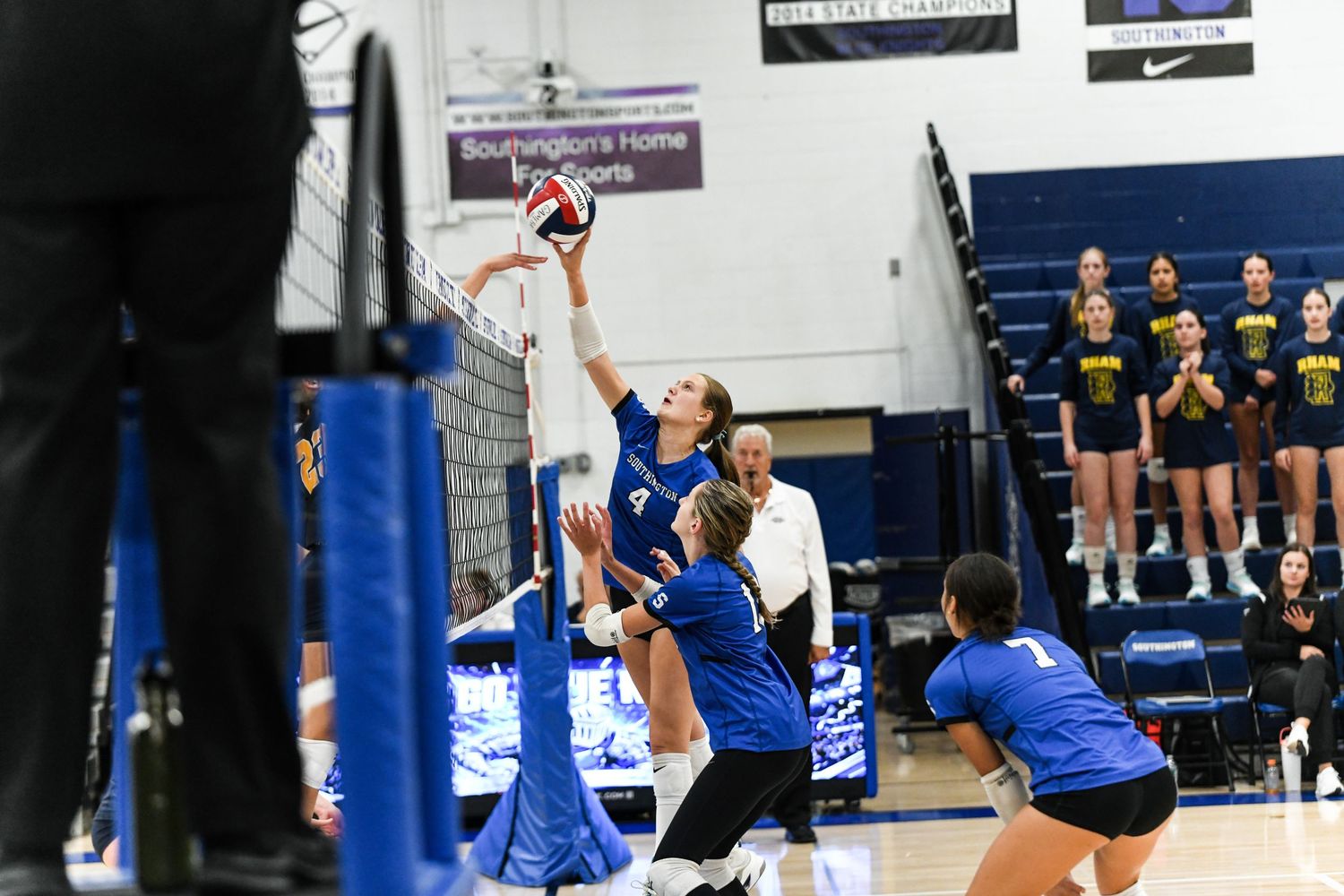 Female volleyball players compete at the net during an indoor match, with one player jumping to spike the ball.