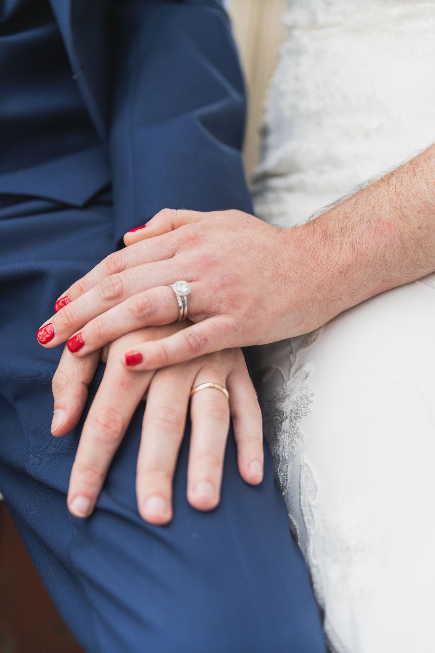 Close-up of newlywed hands with wedding rings against white dress and navy suit fabric.