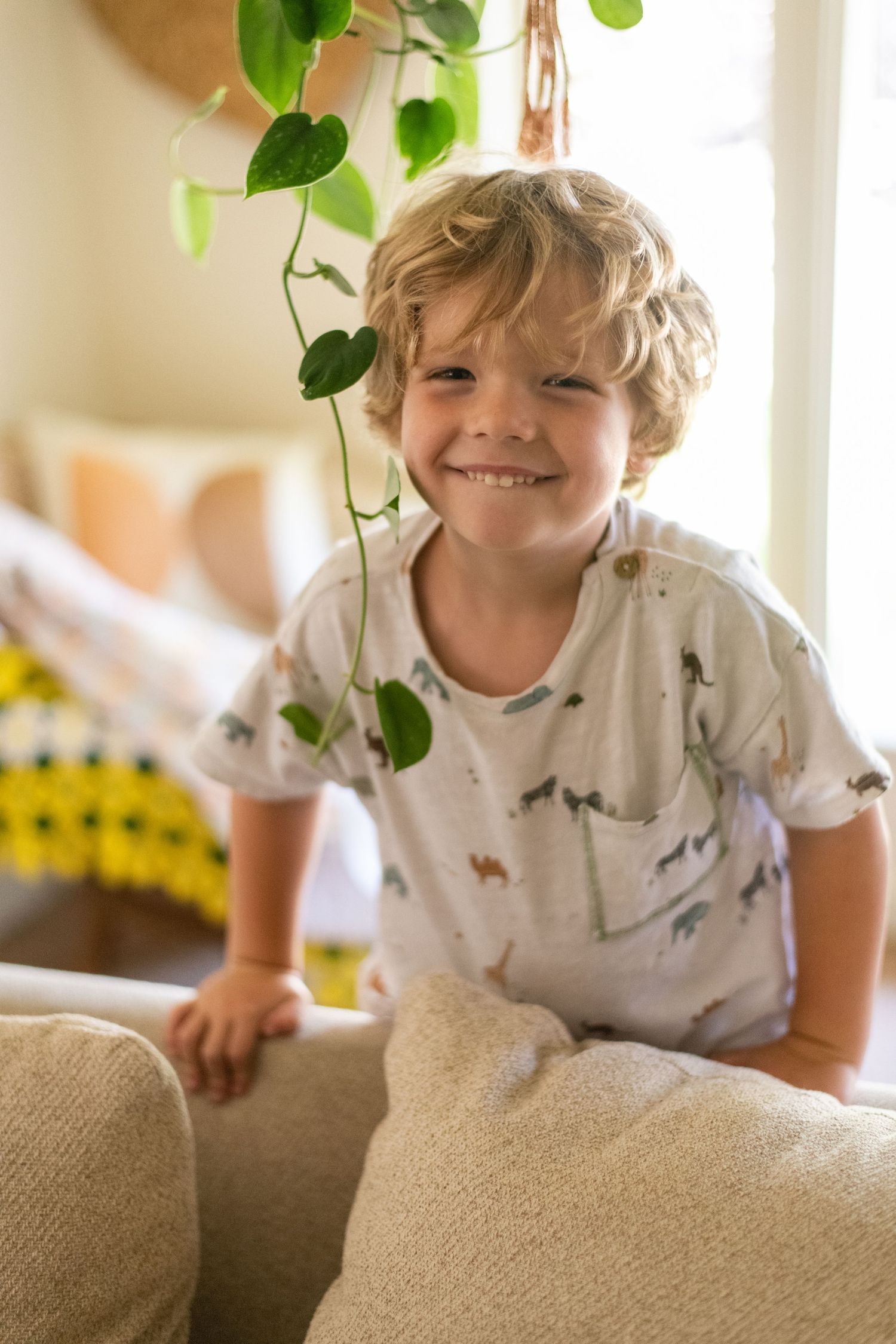 Child in dinosaur print shirt plays energetically on beige couch beneath hanging plants.