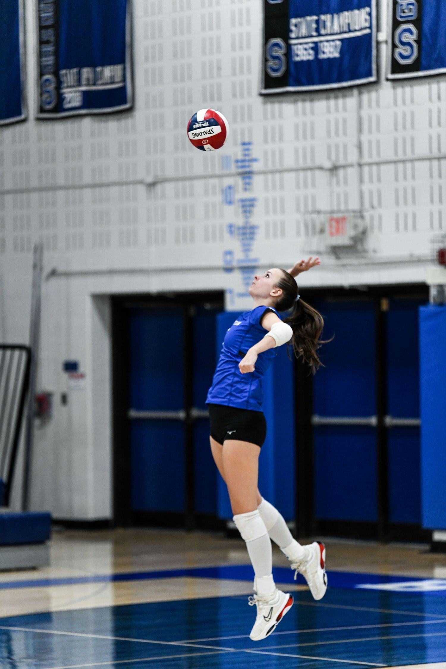 Female volleyball player in blue uniform jumping to serve ball in gymnasium with state championship banners.