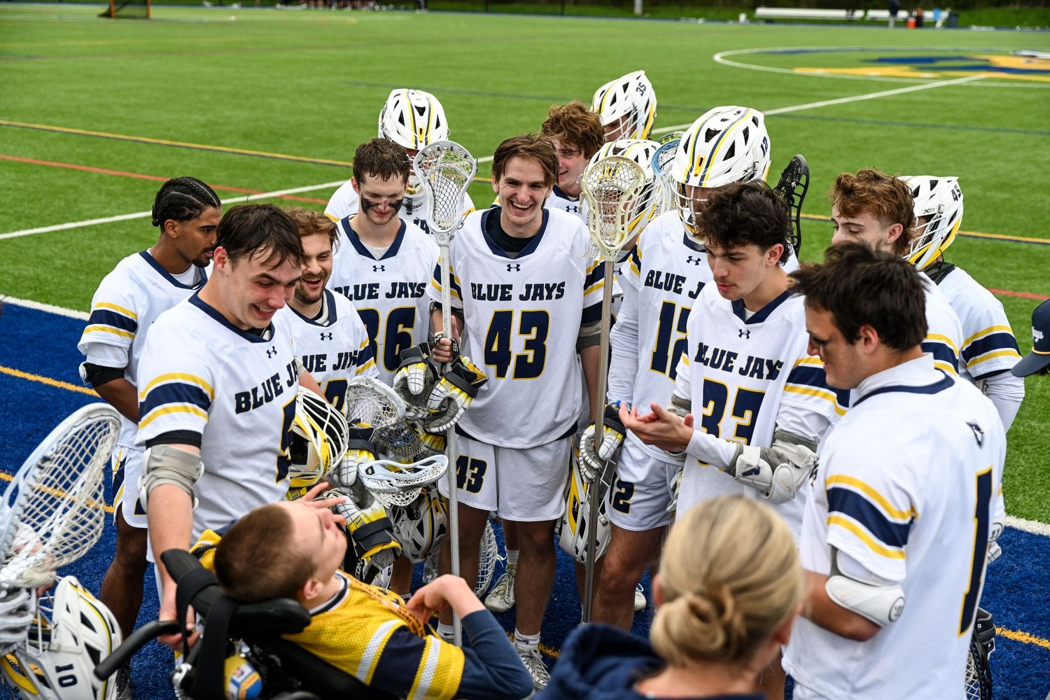 Blue Jays lacrosse team huddles together on field, players in white and navy uniforms celebrating after a game.