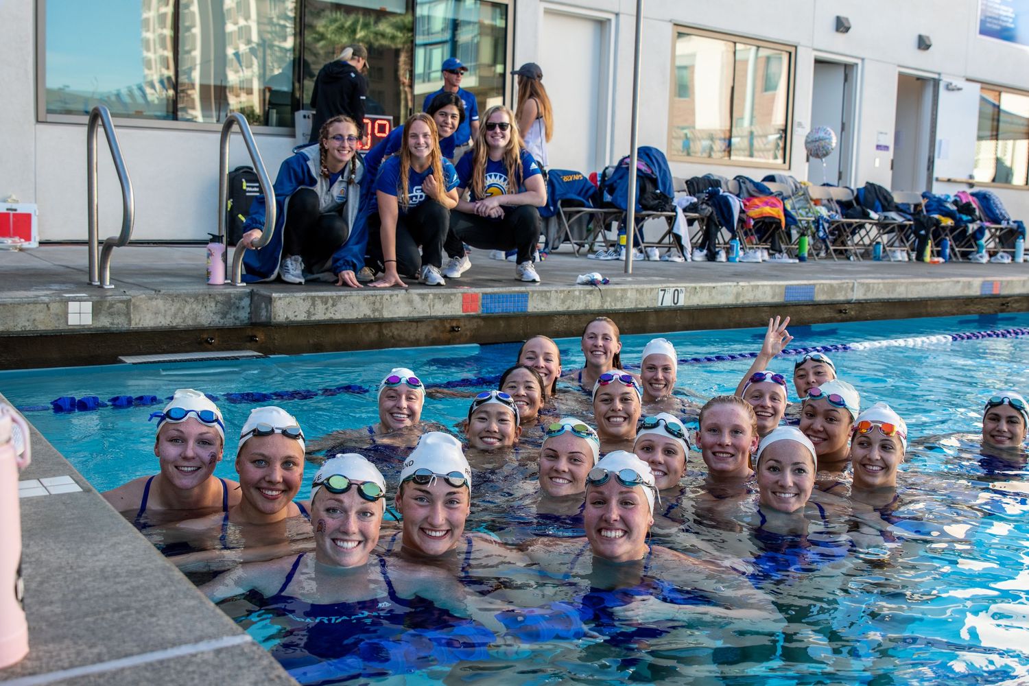 Swim team posing in outdoor pool with coaches on deck, wearing white caps and blue swimsuits.