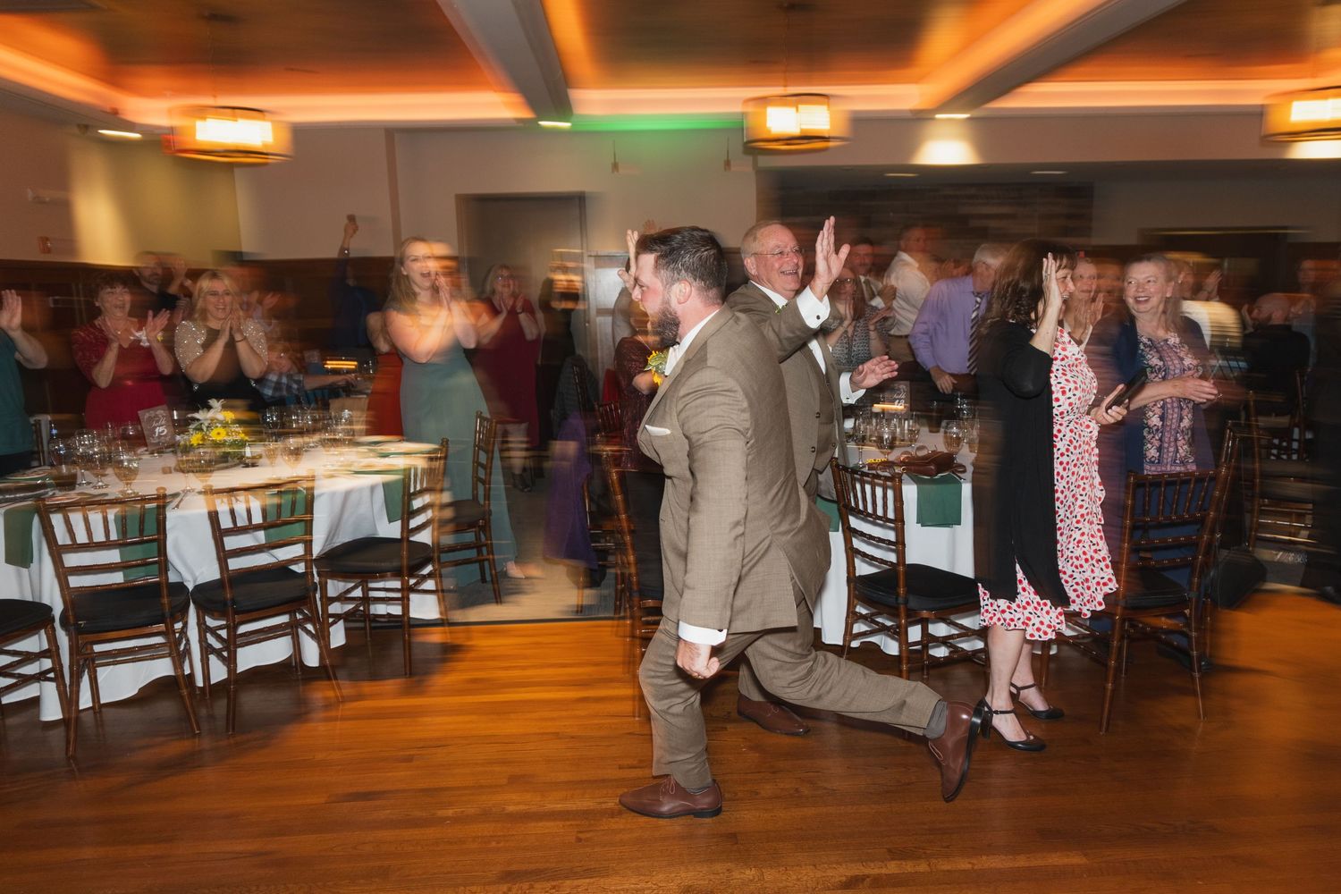 Guests dance and celebrate on a warmly lit wooden dance floor at a wedding reception.