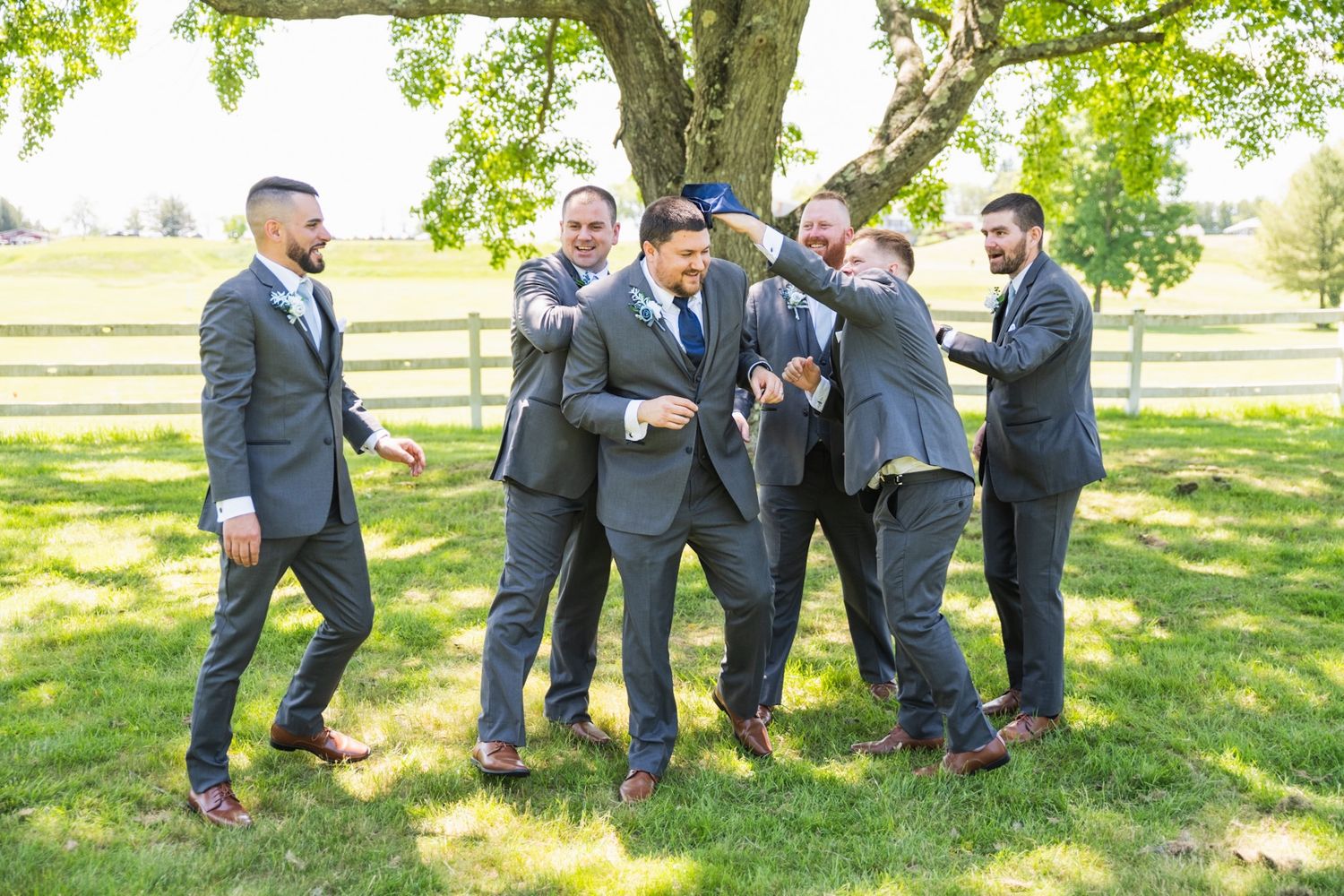Groomsmen in matching gray suits share a lighthearted moment outdoors under a tree.
