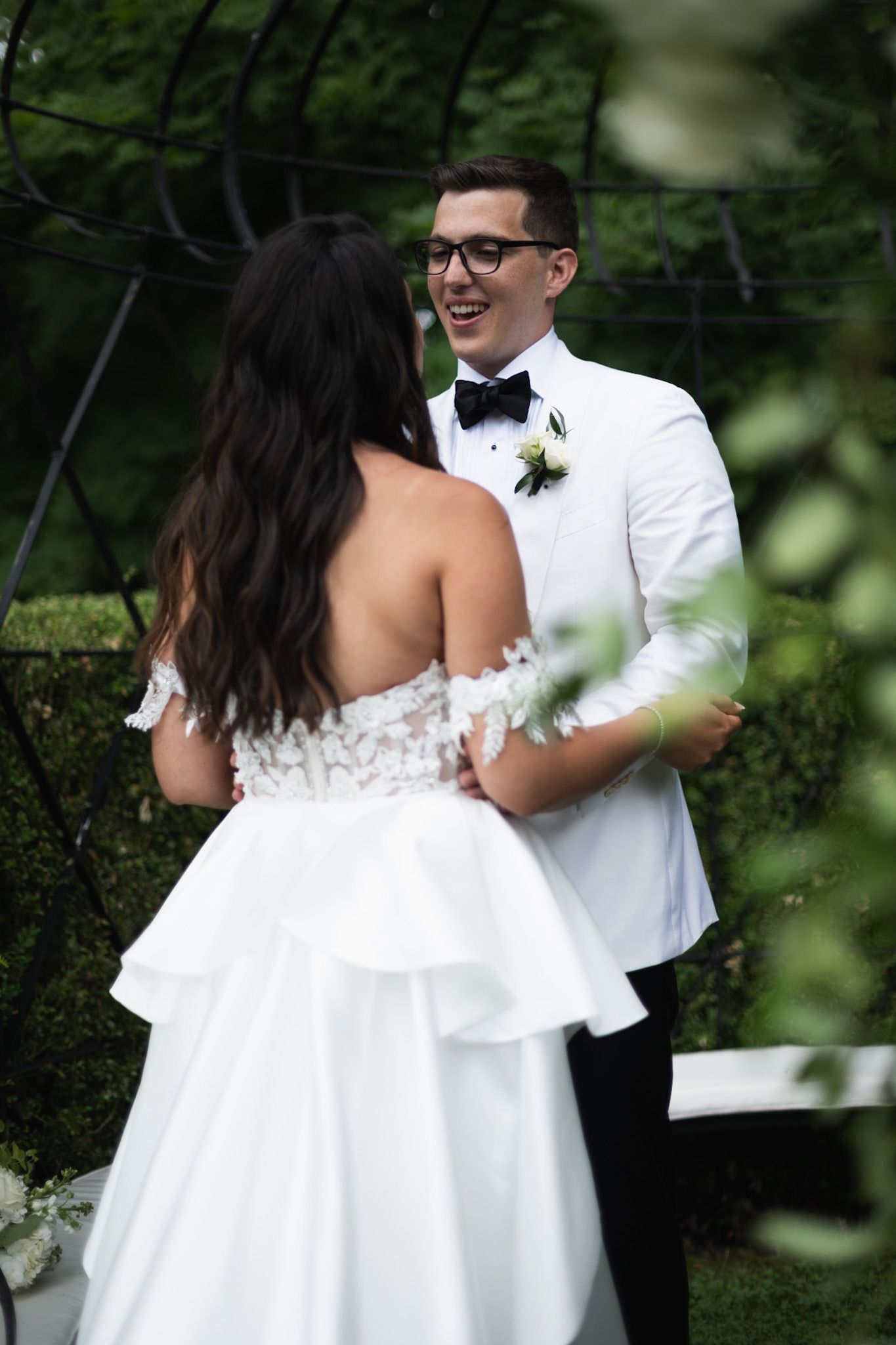 A couple in wedding attire embrace during an outdoor first look moment amid greenery.