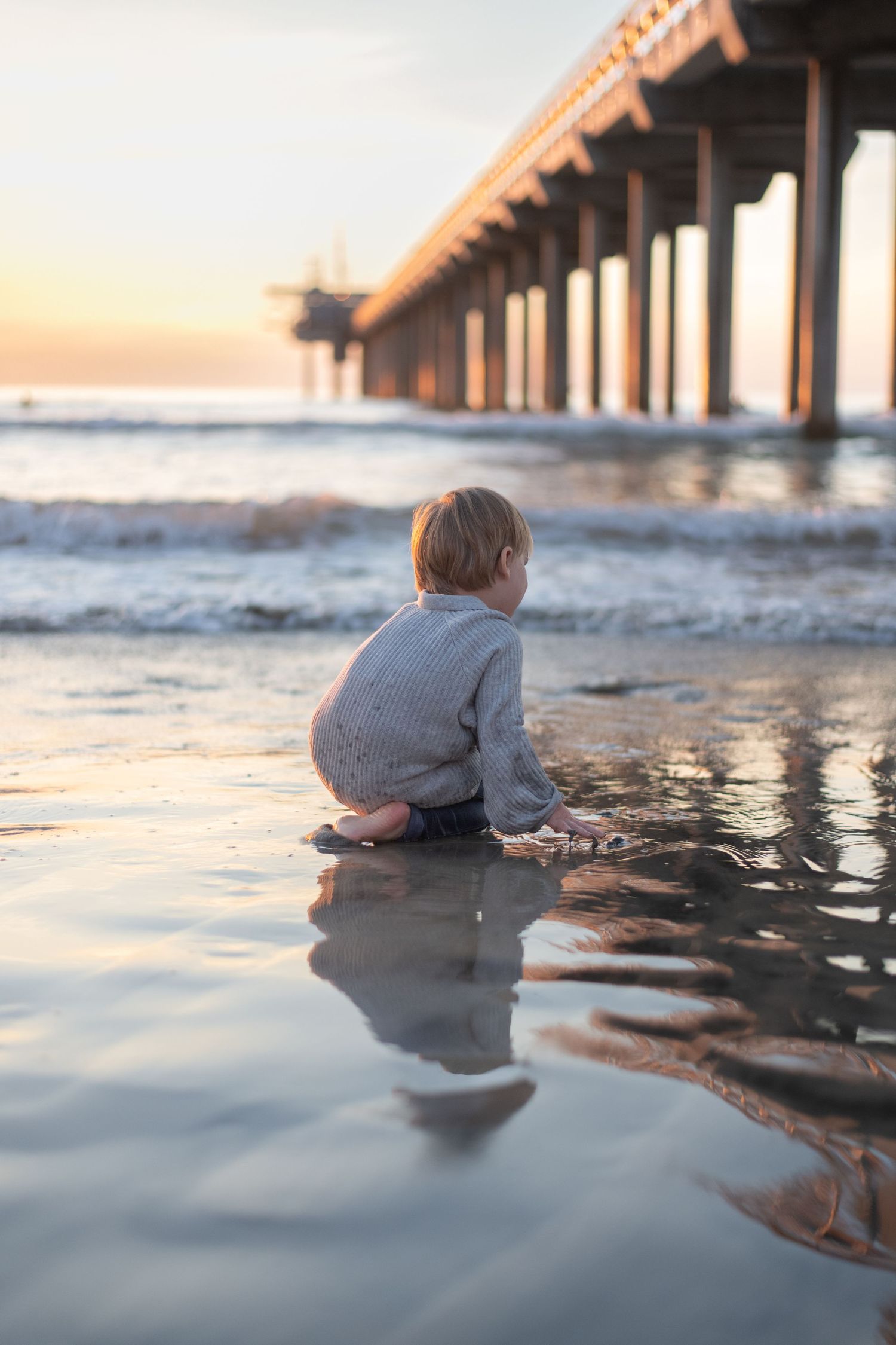 Low-angle beach photograph capturing reflections in wet sand beneath a pier at sunset.