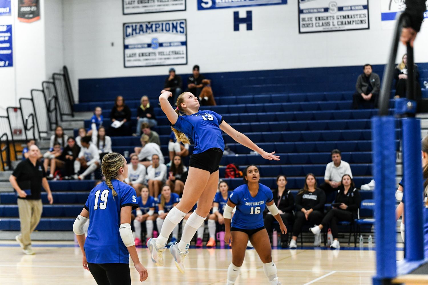 High school volleyball player jumps to spike the ball during an indoor gym match, teammates and crowd visible.