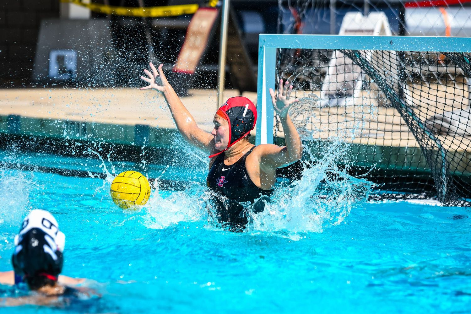 Water polo goalkeeper in red cap leaping to block yellow ball during competitive pool match.