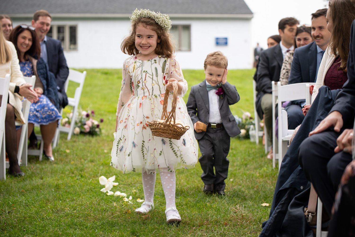 A flower girl in a white dress walks down the aisle during an outdoor ceremony.