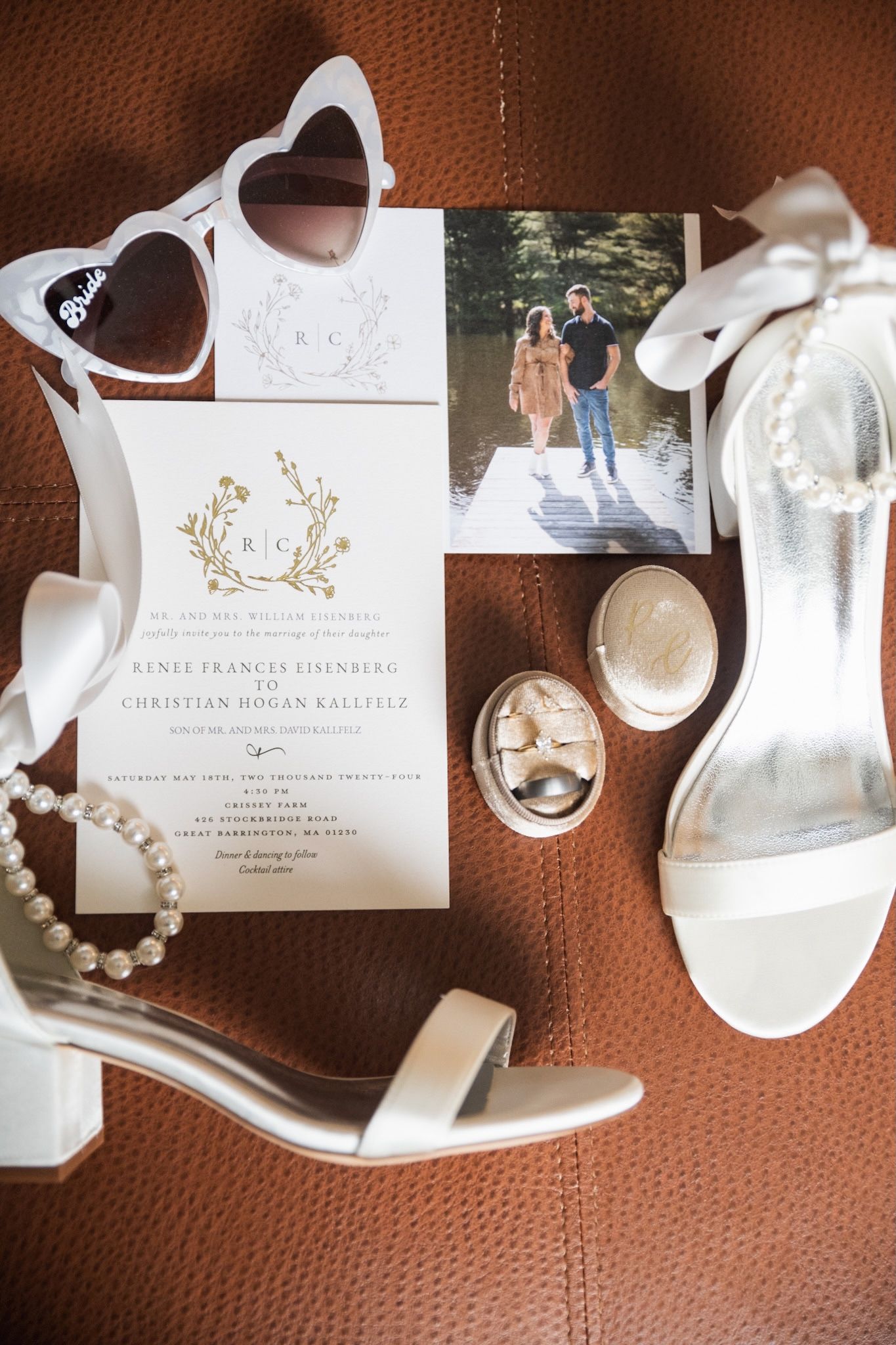 Bridal accessories including white shoes, pearl jewelry and wedding invitations laid out on textured brown leather.