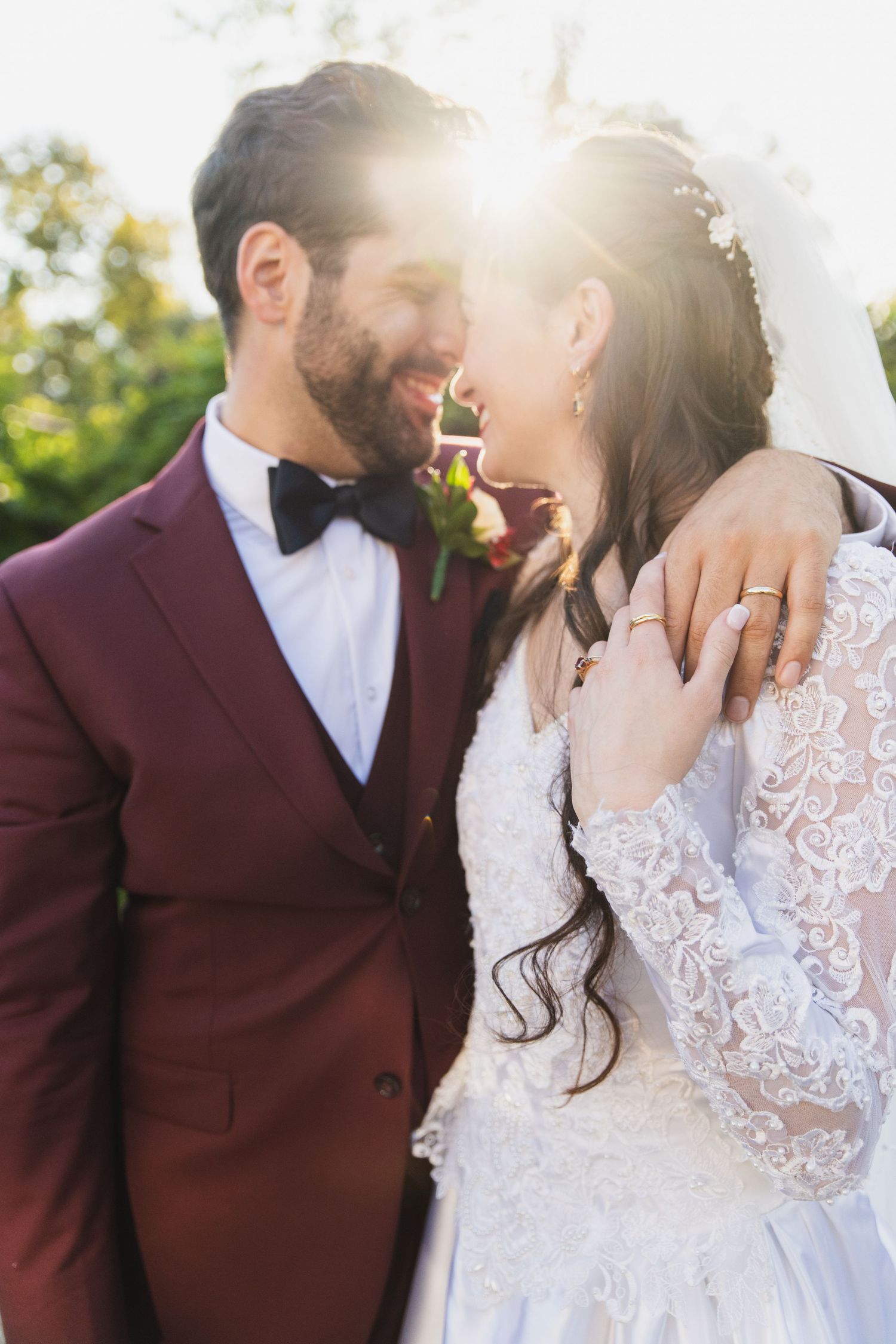 A couple in burgundy suit and white lace dress share an intimate moment in golden sunlight.