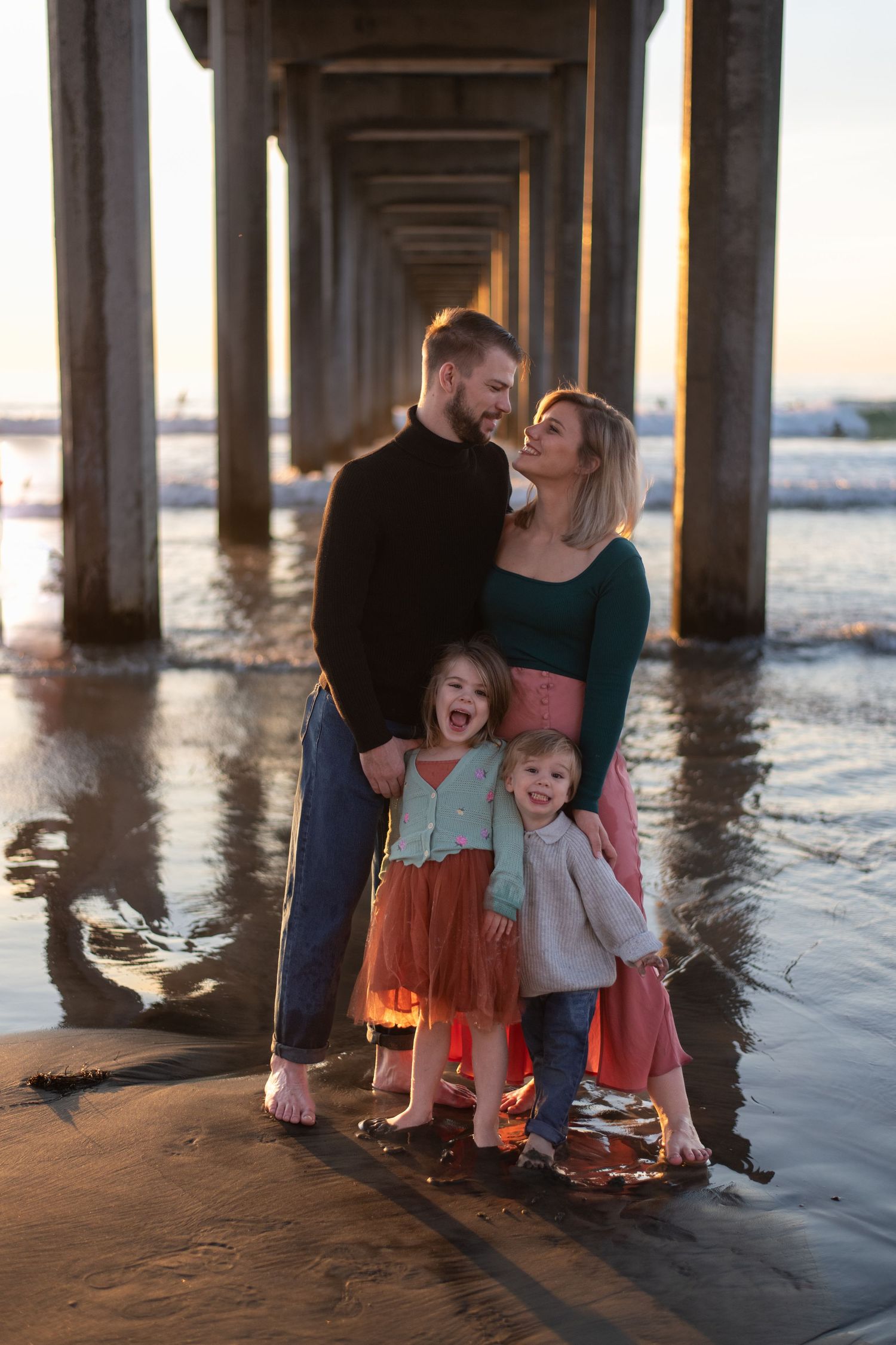 Family playfully interacts under a pier at sunset, creating joyful silhouettes against the ocean waves.
