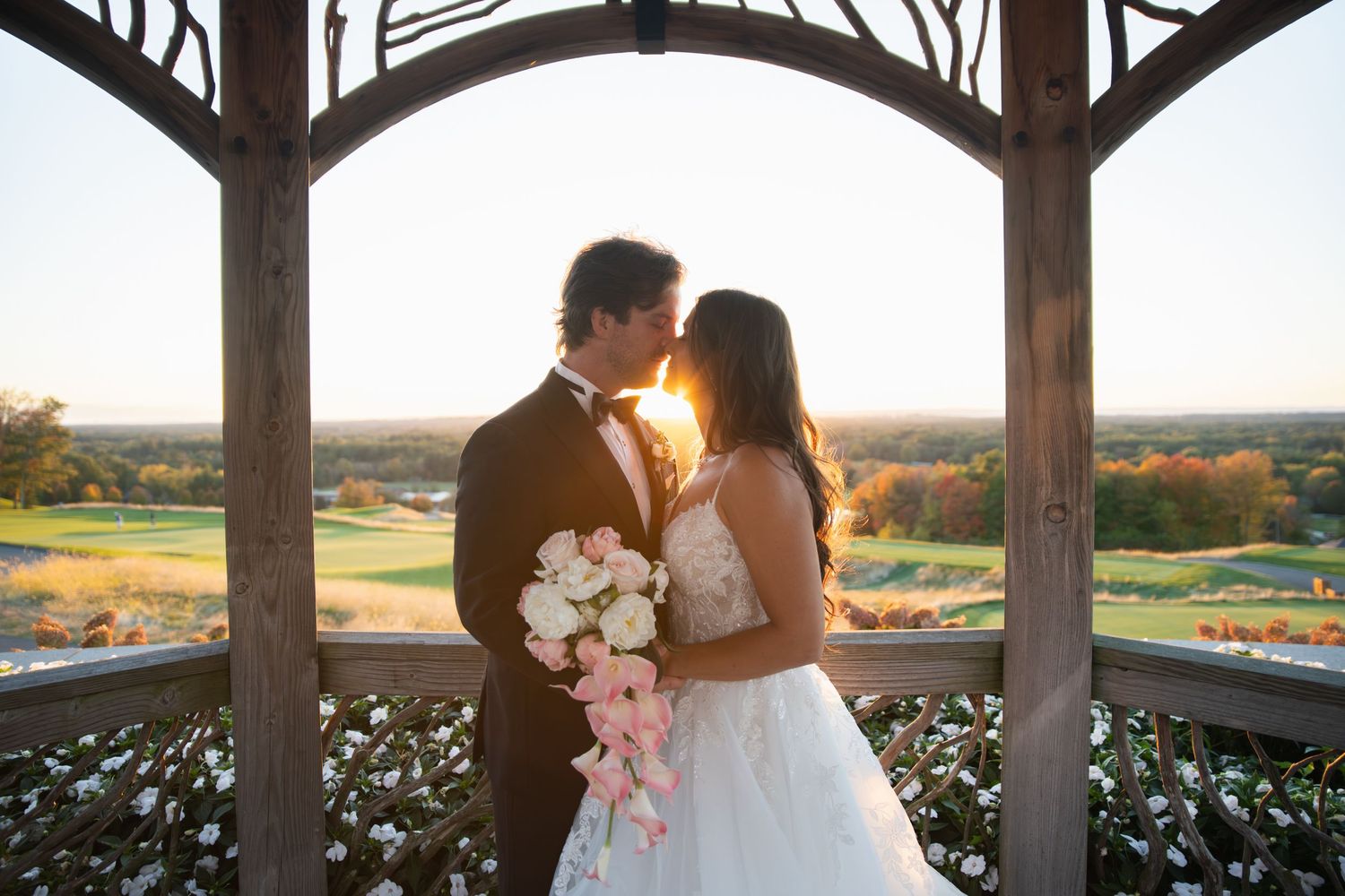 A romantic sunset wedding kiss between a couple in an outdoor gazebo overlooking a scenic landscape.