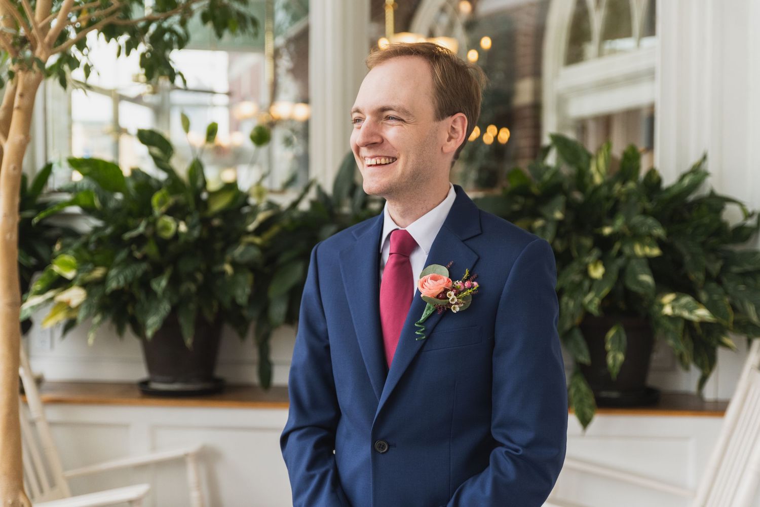 Groomsmen in navy suits and burgundy ties share a laugh during wedding photos near a white porch with greenery.