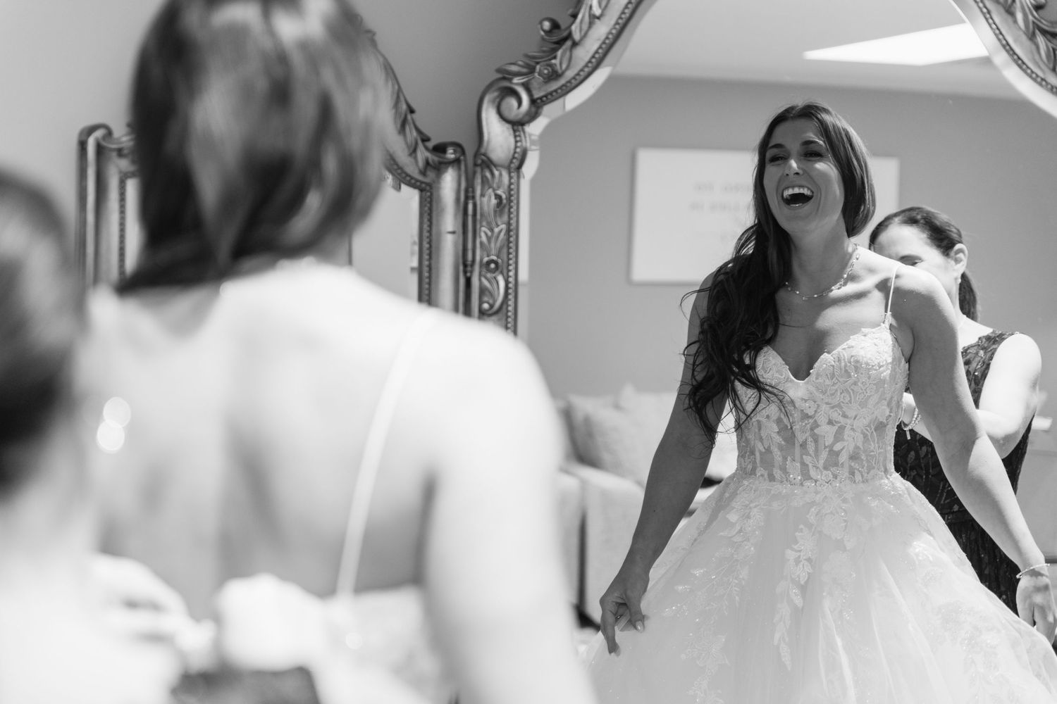 Black and white photo of a bride in a lace wedding dress laughing joyfully while looking at her reflection in a mirror.