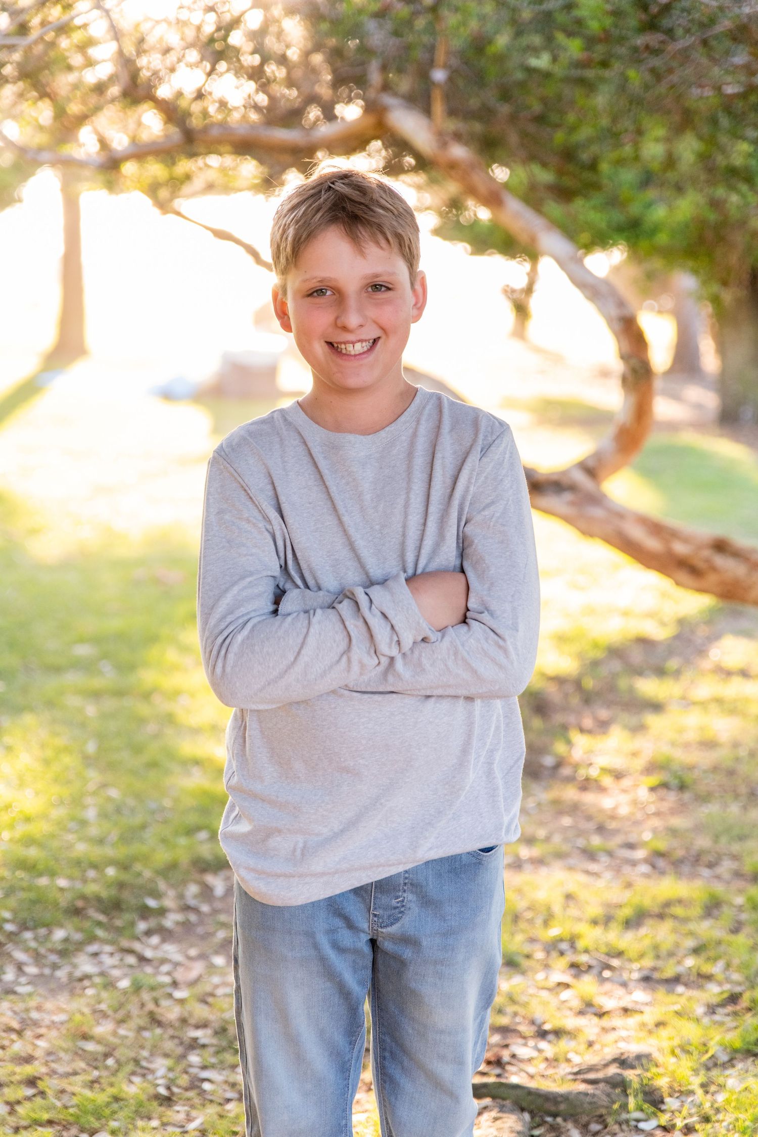 Young boy in gray sweater and jeans posing outdoors with crossed arms.