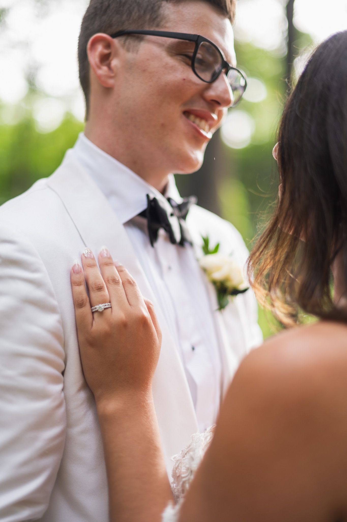 Close-up of groom in white tuxedo and glasses showing wedding ring.