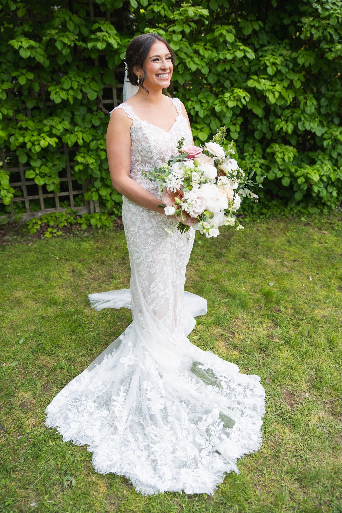 Bride in fitted lace gown twirls while holding white bouquet on green lawn.