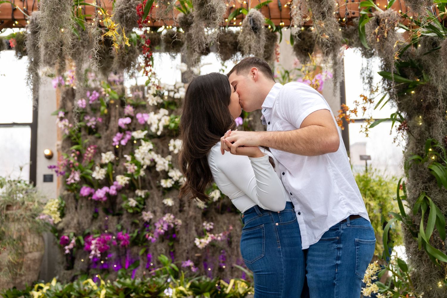 A couple shares a kiss under a beautiful floral arch with purple flowers.