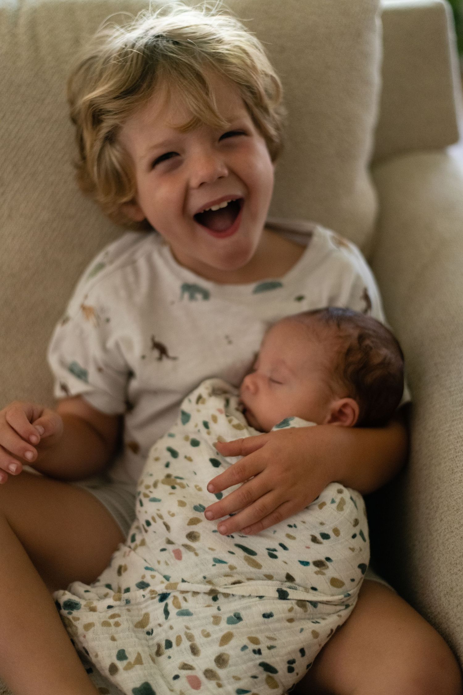 Siblings sharing sweet moment on couch with newborn baby wrapped in patterned swaddle.