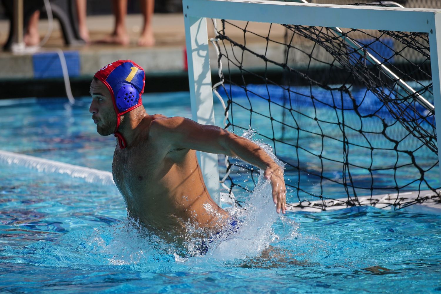 Water polo goalkeeper defends goal net in outdoor pool during competitive water polo match.