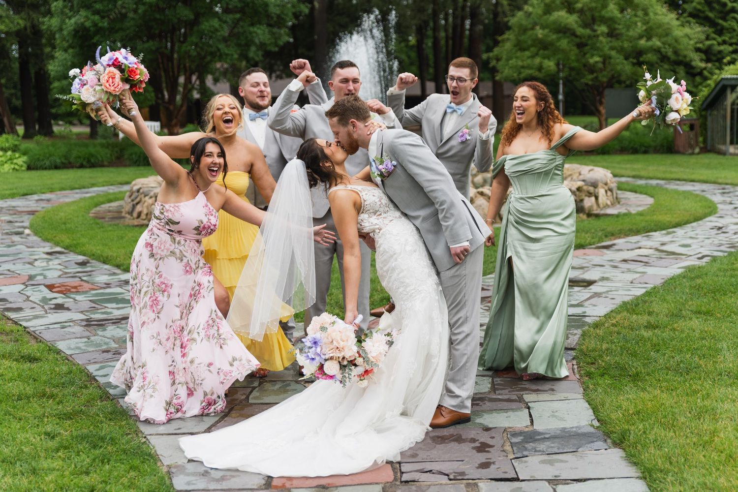 Wedding party poses playfully on wet stone pathway during outdoor celebration.